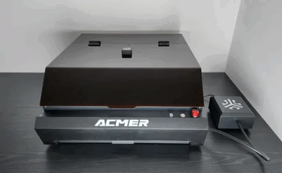 Acmer P3 24W enclosed laser engraver with protective lid and external control module on a black desk