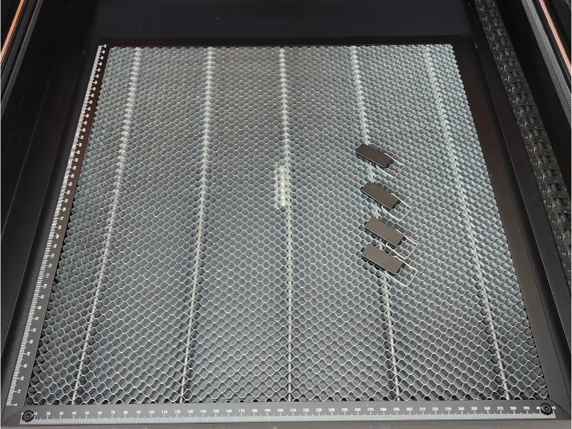 A laser cutting machine bed featuring a honeycomb grid pattern with four small rectangular metal pieces placed on its right side. Measurement markings are visible along the edges.