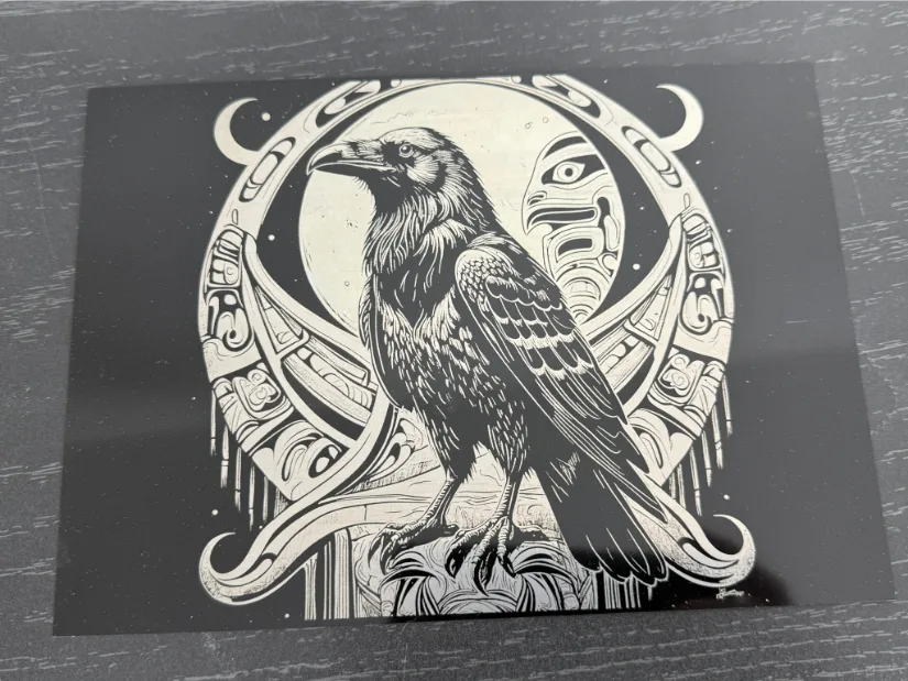 A detailed illustration of a raven perched on a rock. The raven's realistic feathers and sharp gaze are framed by a circular design of abstract tribal elements—including crescent moons and faces—with a luminous full moon in the background.
