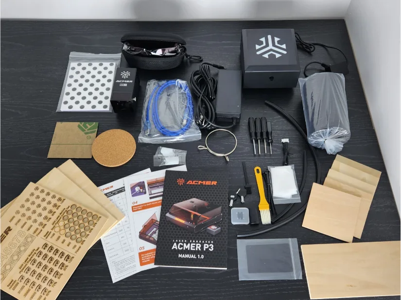 Unboxed components of the Acmer P3 24W laser engraver kit laid out on a black surface, including cables, controller, laser module, sample materials, and user manual