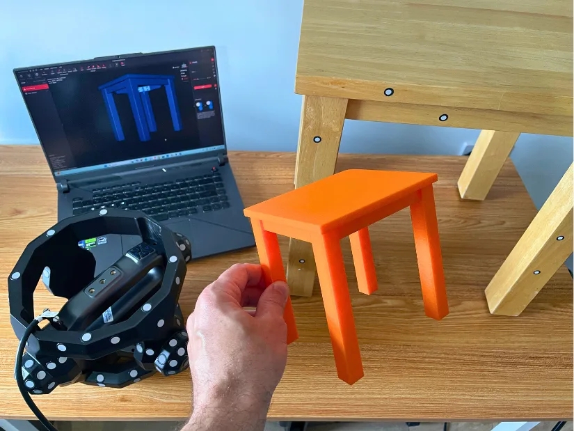 3D printed orange model of a small table held in front of the original, with scanner, laptop showing mesh, and markers still on the full-size furniture