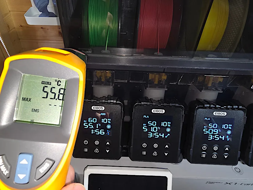 Infrared thermometer displaying 55.8°C held near an EIBOS Series X Tetras dryer module, verifying heating accuracy against digital screen readings on three active drying bays.
