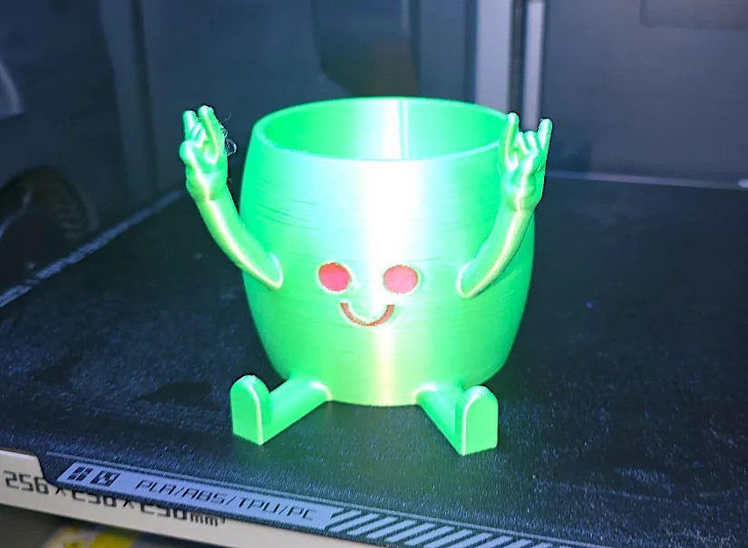 Green 3D printed character pot with red eyes and a smiling face, sitting with raised hands in a “rock on” gesture, printed as a multi-color test using dried filament.