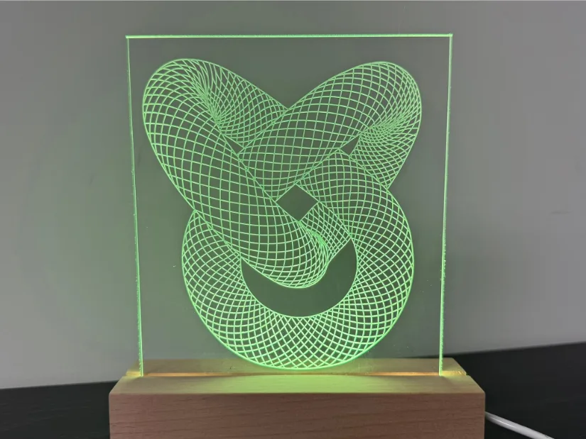 Edge‑lit acrylic lamp engraved with an intricate torus‑knot grid pattern using a 30‑degree V‑bit on the Lunyee 3018 Pro Ultra CNC router, glowing green in a beech wood LED base.