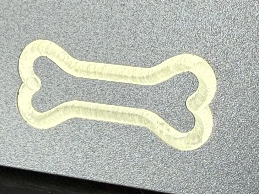 Close‑up of a dog‑bone outline milled into an anodized aluminum plate, tool‑marked by a 1/8‑inch tungsten‑carbide end mill on the Lunyee 3018 Pro Ultra CNC router.