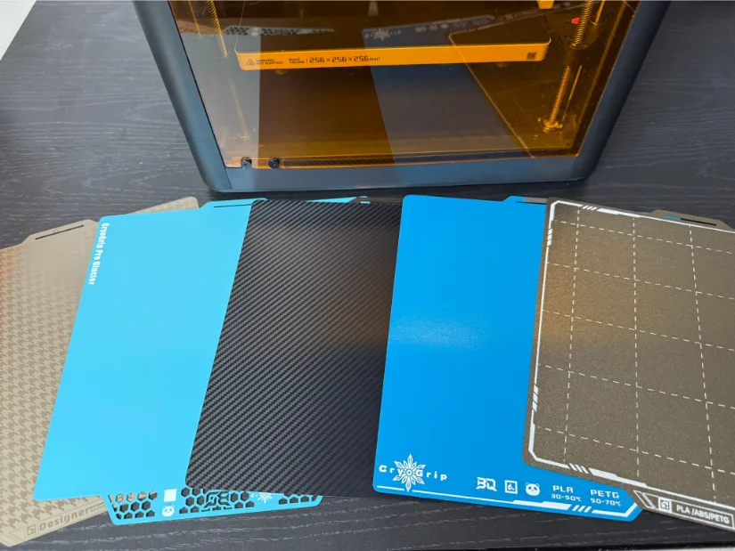 Close-up of five BIGTREETECH Panda magnetic build plates for Bambu Lab 3D printers—houndstooth designer plate, smooth turquoise PEI, carbon-fiber textured plate, CryoGrip cold plate, and textured PEI grid—fanned out in front of an enclosed Bambu Lab printer.