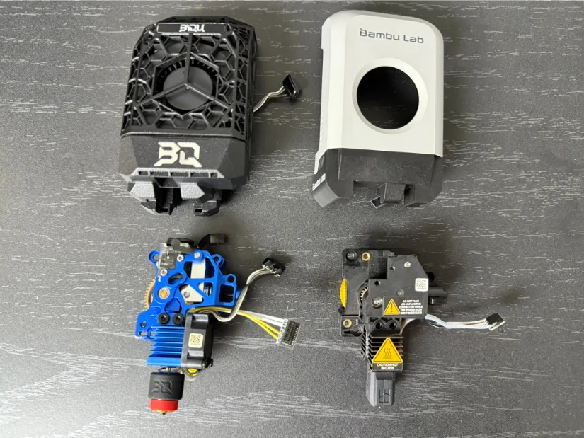 Side-by-side comparison of four Bambu-compatible tool-head parts on a dark desk: the lattice-style BIGTREETECH JetDock cover and blue Chain all-metal extruder with Volcano-type hotend (left) next to the grey stock Bambu Lab tool-head housing and factory extruder/hotend assembly (right).
