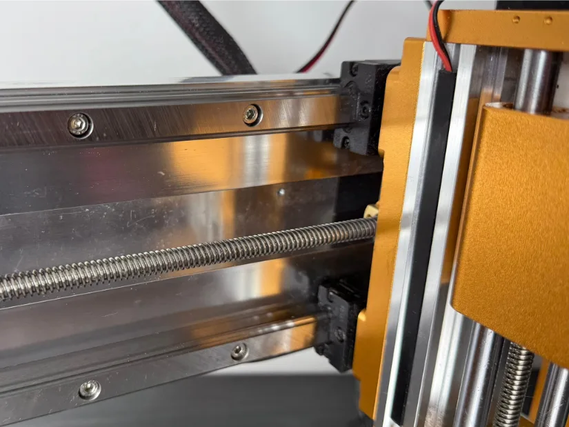 Close‑up of the Lunyee 3018 Pro Ultra’s X‑axis: hardened steel linear guide rails with recirculating‑ball blocks and a stainless lead screw driving the gold‑anodized Z‑axis carriage.