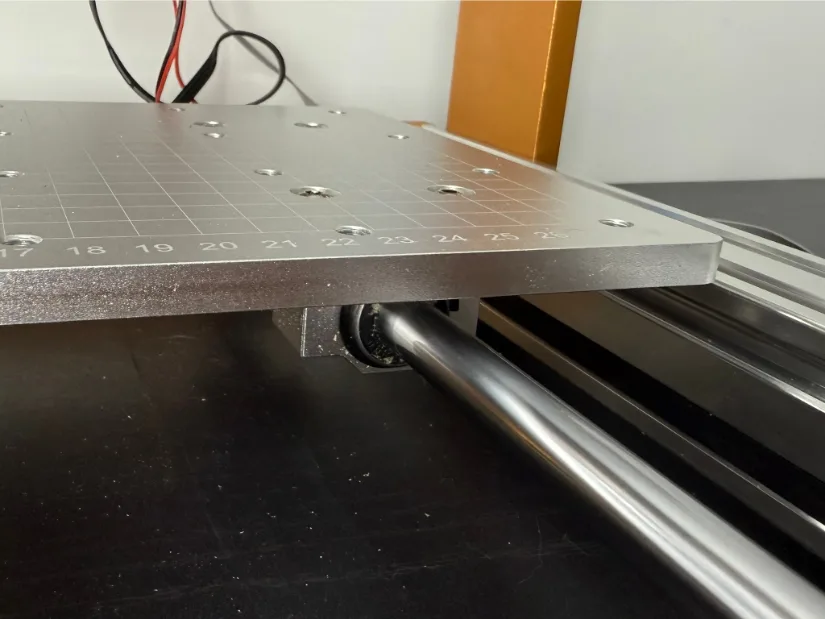 Close‑up of the Lunyee 3018 Pro Ultra’s Y‑axis: steel linear guide rod and bearing block supporting the aluminum T‑slot work bed with engraved grid markings.
