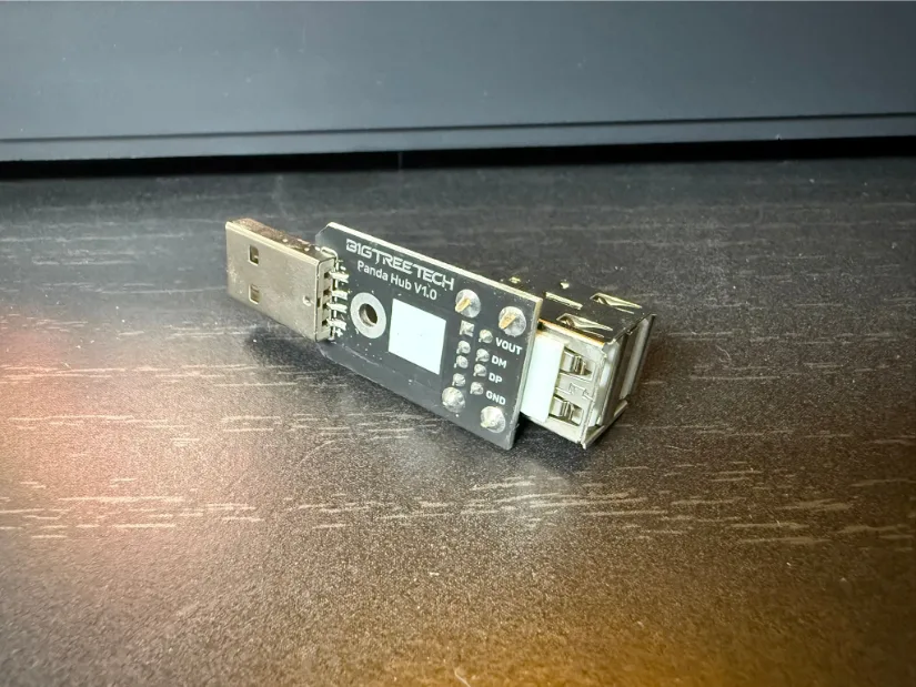 BIGTREETECH Panda Hub V1.0 inline USB board with a male USB-A plug, stacked dual USB-A ports, pin header, and labeled test pads resting on a black tabletop.