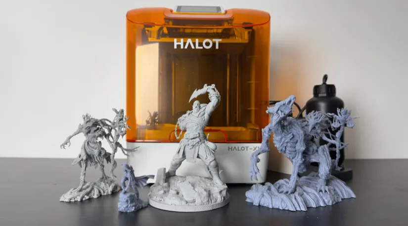 Piocreat HALOT-X1 resin 3D printer with orange cover and Auto Feed Unit, shown with finished test prints—Wicked3D Kratos and Nerikson Famine Horseman miniatures—on a desktop