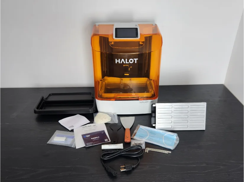 Piocreat HALOT-X1 resin 3D printer unboxed with accessories: quick-release build plate with removal tabs, print tray, metal and plastic scrapers, power cable, hex keys, masks, gloves, lubricant, and paperwork on a black table