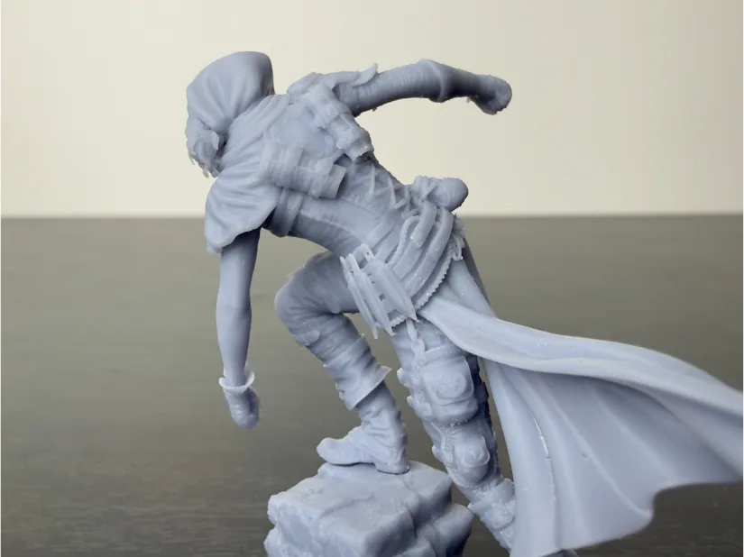 Rear/side view of Nerikson Enika the Assassin 75 mm resin miniature—unpainted gray—with flowing cape, straps and pouches, standing on a rock, printed on Piocreat HALOT-X1