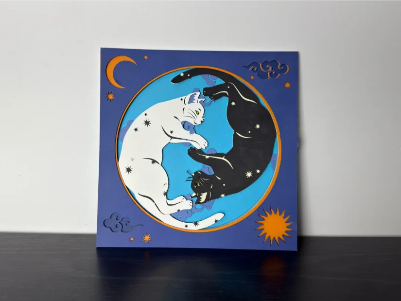 Assembled nine-layer paper cut made with the xTool M1 Ultra, showing black and white cats curled in a yin-yang pose inside a circular frame with moons, stars, and clouds.
