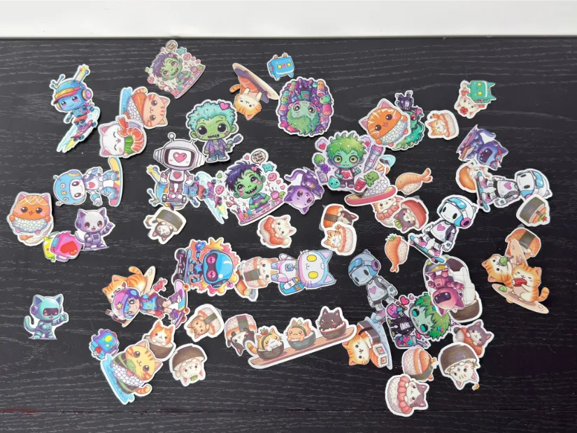 Assorted cute character stickers—robots, cats, zombies, and food-themed designs—printed and kiss-cut with the xTool M1 Ultra, scattered on a black tabletop.