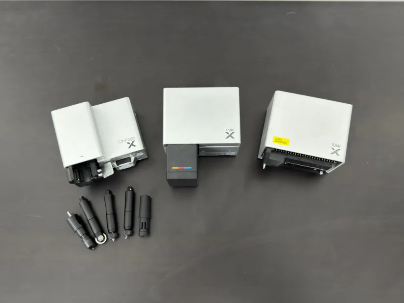 xTool M1 Ultra modules on a table: Multi-Function Carriage (left), Inkjet module (center), 10W laser module (right), with four tool heads in front (pen holder, cutting blade, rotary blade, and foil tip).xTool M1 Ultra modules on a table: Multi-Function Carriage (left), Inkjet module (center), 10W laser module (right), with four tool heads in front (pen holder, cutting blade, rotary blade, and foil tip).
