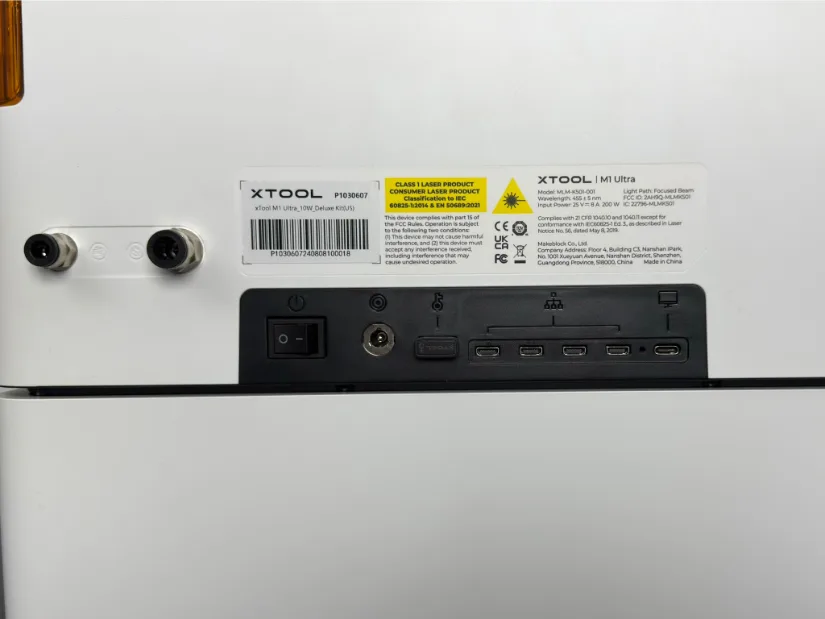 Rear panel of the xTool M1 Ultra showing compliance labels, power switch, DC power jack, reset/service pinhole, “TOOLS” port, four USB-C ports, one USB-A port, and hose fittings on the left.
