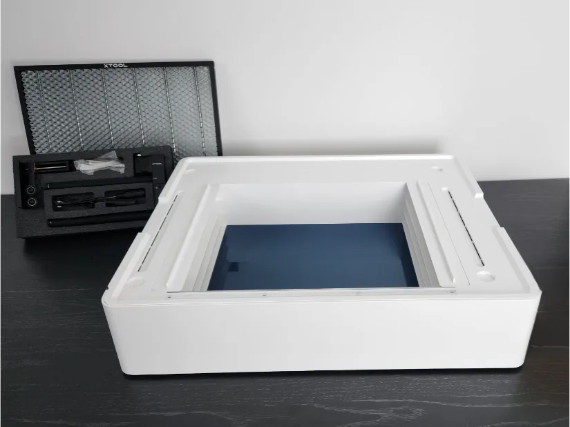 xTool M1 Ultra Riser Base on a table, with the honeycomb panel and accessory kit (legs and hardware) shown in the background.