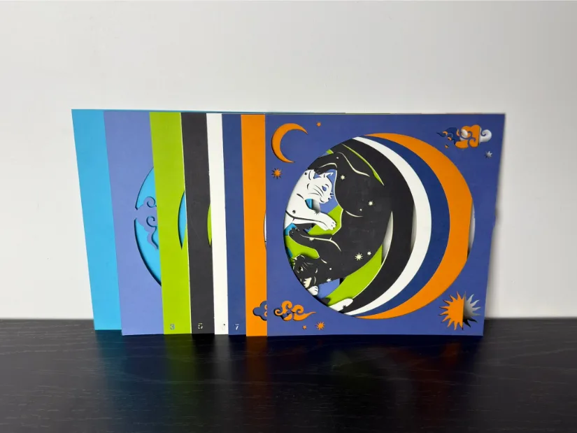 Multi-layer paper artwork cut with the xTool M1 Ultra, featuring a sleeping cat within crescent moon shapes and night-sky motifs; individual colored layers (blue, green, black, white, orange) are shown.