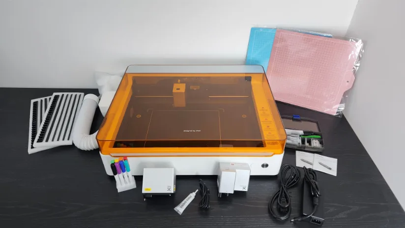 xTool M1 Ultra on a table with amber lid, surrounded by included accessories—exhaust hose and vent grills, sticky cutting mats (blue and pink), pen set, modules, power supplies, cables, and small toolkit items.