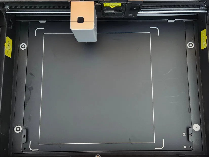 Inside view of the xTool M1 Ultra showing the carriage over the flat work bed with white alignment marks, magnetic anchors, and safety labels along the sides.