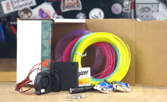Print kit contents including colorful 3D printing filament, electronics, fan, battery, and hardware components laid out on a workbench