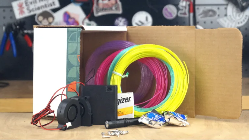 Print kit contents including colorful 3D printing filament, electronics, fan, battery, and hardware components laid out on a workbench