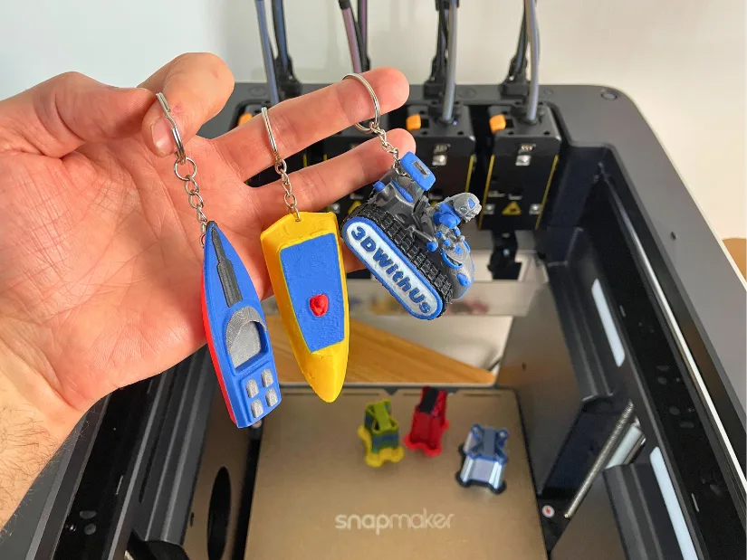 3D printed keychain miniatures from scans including RC boats and DeathRacer displayed in front of Snapmaker U1