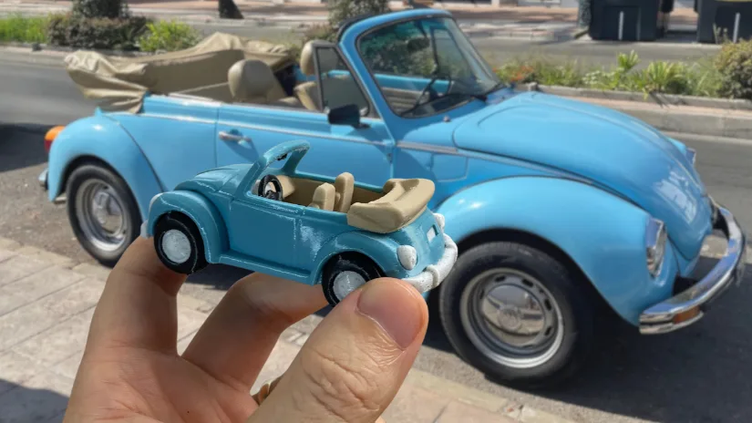 3D printed VW Beetle miniature created with Meshy AI held in front of the original car